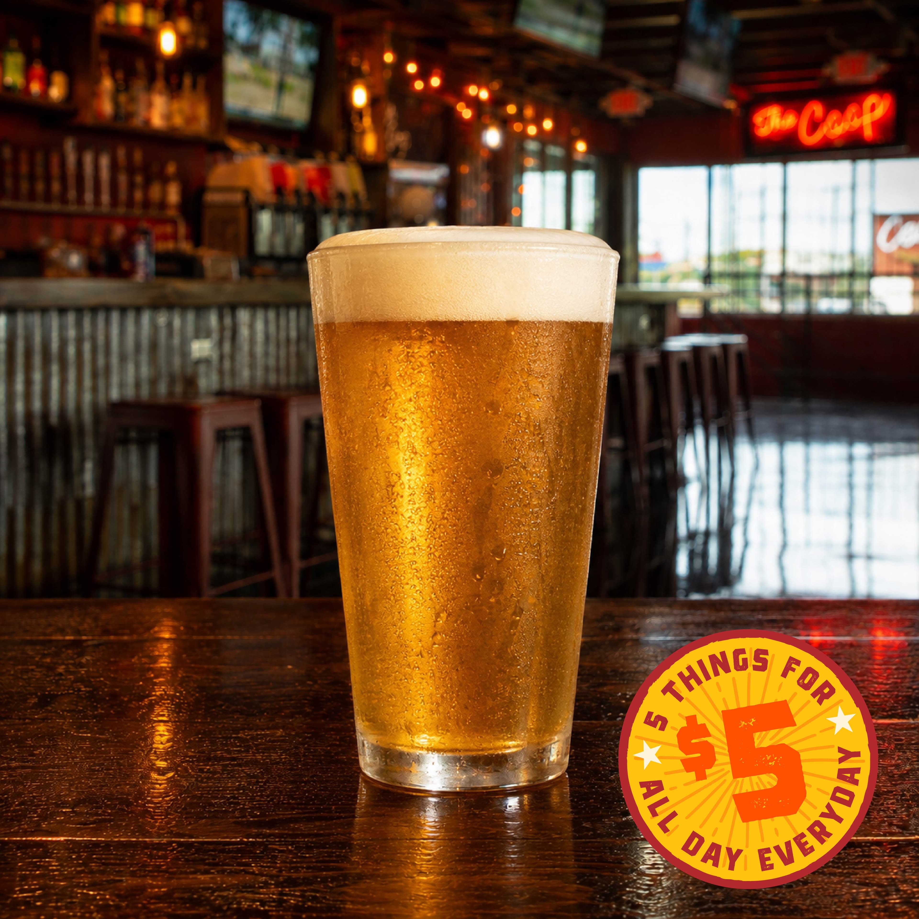 PF HappyHour Image 04 A frosty pint of beer sits on a bar counter in a cozy, dimly lit bar with a 'The Coop' neon sign in the background.
