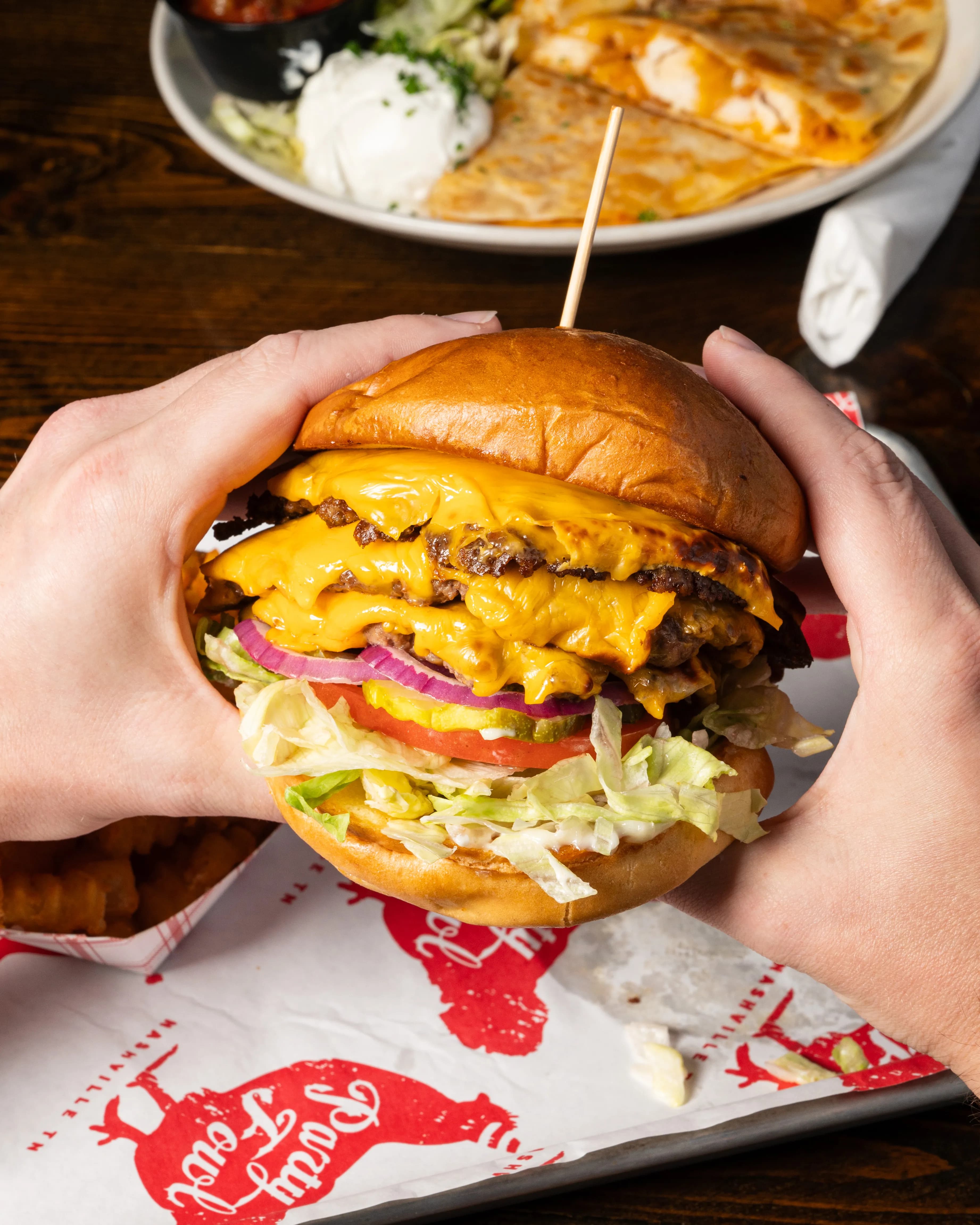 Tripledogdaresmashburger A hand holding a towering triple cheeseburger with layers of beef, melted cheese, lettuce, tomato, and red onion.
