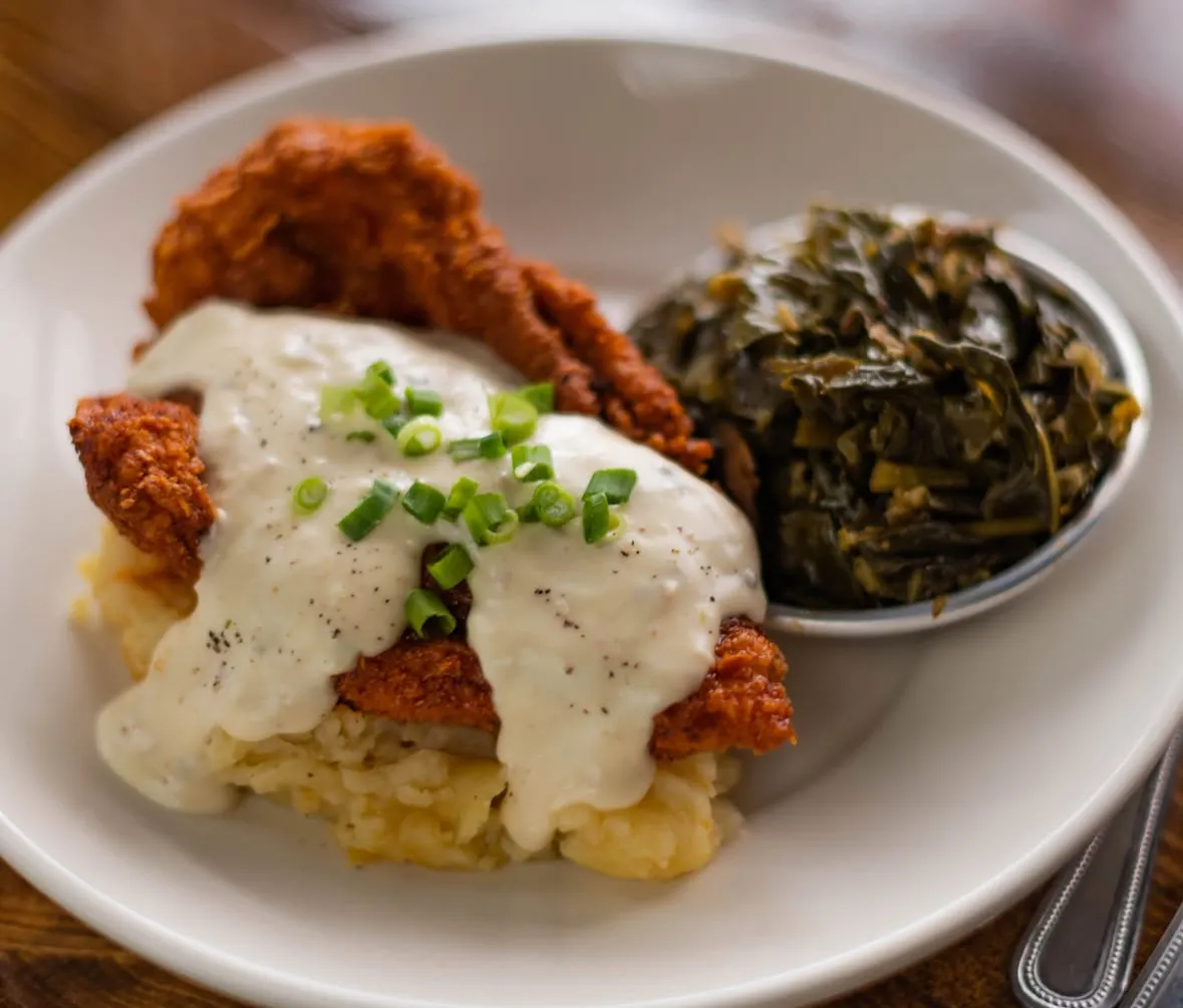 Chickenfriedchicken A plate featuring fried chicken topped with creamy gravy, served with mashed potatoes and collard greens.