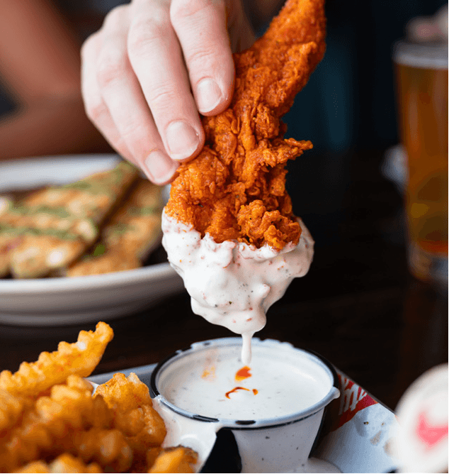 Hot Chicken A hand dipping a crispy fried chicken wing into creamy sauce, surrounded by other fried foods and a drink.